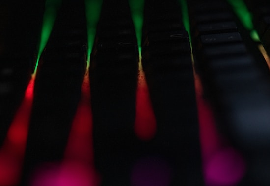 A keyboard with backlit RGB lighting creates a dramatic effect with colors blending from green to red. The lighting casts colorful glows between the dark keys, producing a vivid and dynamic appearance.