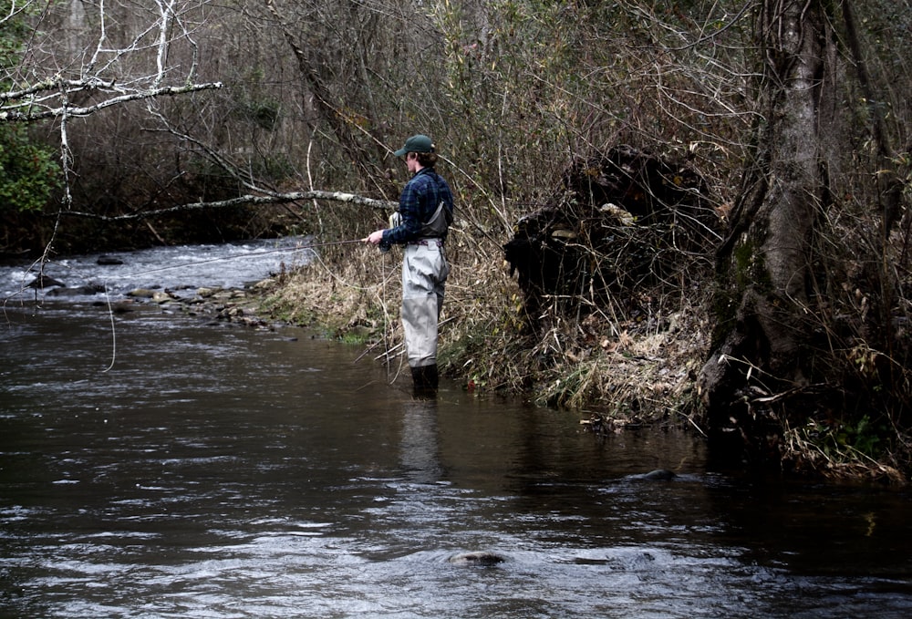 man in blue jacket and gray pants standing on brown rock in river during daytime