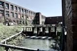 An industrial setting featuring a red brick building with multiple windows, surrounded by overgrown vegetation. In the foreground, there is a water control structure with flowing water beneath it, with metal pipes extending over a grassy area. The scene conveys a blend of natural and man-made elements.