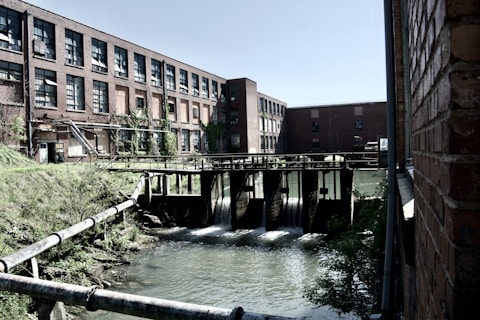 An industrial setting featuring a red brick building with multiple windows, surrounded by overgrown vegetation. In the foreground, there is a water control structure with flowing water beneath it, with metal pipes extending over a grassy area. The scene conveys a blend of natural and man-made elements.