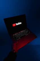 A person holds an open laptop with a dark screen displaying the YouTube logo in bright red and white. The surroundings are enveloped in a blue hue, while the light from the screen casts a red glow on the person's hand and wrist.