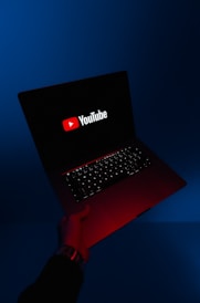 A person holds an open laptop with a dark screen displaying the YouTube logo in bright red and white. The surroundings are enveloped in a blue hue, while the light from the screen casts a red glow on the person's hand and wrist.