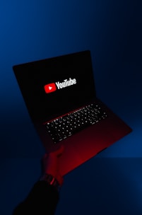 A person holds an open laptop with a dark screen displaying the YouTube logo in bright red and white. The surroundings are enveloped in a blue hue, while the light from the screen casts a red glow on the person's hand and wrist.