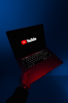 A person holds an open laptop with a dark screen displaying the YouTube logo in bright red and white. The surroundings are enveloped in a blue hue, while the light from the screen casts a red glow on the person's hand and wrist.