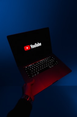 A person holds an open laptop with a dark screen displaying the YouTube logo in bright red and white. The surroundings are enveloped in a blue hue, while the light from the screen casts a red glow on the person's hand and wrist.
