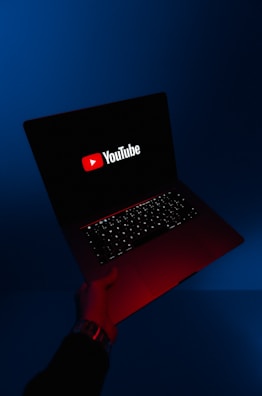 A person holds an open laptop with a dark screen displaying the YouTube logo in bright red and white. The surroundings are enveloped in a blue hue, while the light from the screen casts a red glow on the person's hand and wrist.