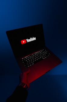 A person holds an open laptop with a dark screen displaying the YouTube logo in bright red and white. The surroundings are enveloped in a blue hue, while the light from the screen casts a red glow on the person's hand and wrist.