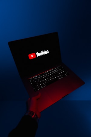 A person holds an open laptop with a dark screen displaying the YouTube logo in bright red and white. The surroundings are enveloped in a blue hue, while the light from the screen casts a red glow on the person's hand and wrist.