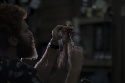 A person with curly hair and a beard is cutting a section of long hair using scissors. The scene is dimly lit, creating a moody atmosphere with dark shadows. The background is blurred, showing hints of shelves and indistinct objects.