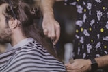 A person is having their hair cut. The barber is holding a pair of scissors and a comb, focusing on trimming the hair. The customer is draped with a striped barber cape, and the barber is wearing a shirt with a space-themed pattern.