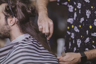 A person is having their hair cut. The barber is holding a pair of scissors and a comb, focusing on trimming the hair. The customer is draped with a striped barber cape, and the barber is wearing a shirt with a space-themed pattern.