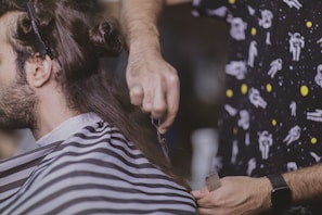 A person is having their hair cut. The barber is holding a pair of scissors and a comb, focusing on trimming the hair. The customer is draped with a striped barber cape, and the barber is wearing a shirt with a space-themed pattern.