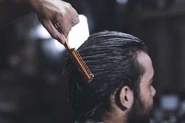 A close-up of a person with dark hair, sitting in a salon chair. Their hair appears damp and is being combed back with a wide-toothed orange comb by a hand visible at the top of the image. The background is blurred, focusing attention on the grooming process.