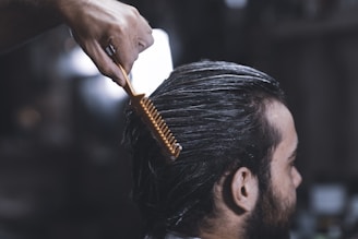 A close-up of a person with dark hair, sitting in a salon chair. Their hair appears damp and is being combed back with a wide-toothed orange comb by a hand visible at the top of the image. The background is blurred, focusing attention on the grooming process.