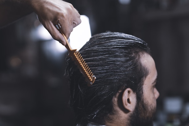 A close-up of a person with dark hair, sitting in a salon chair. Their hair appears damp and is being combed back with a wide-toothed orange comb by a hand visible at the top of the image. The background is blurred, focusing attention on the grooming process.