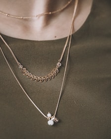 A close-up view of a person wearing multiple delicate necklaces. The jewelry consists of thin gold chains, featuring a detailed leaf-like pattern and small round pendants. The background is a green fabric or clothing, which complements the gold tones of the necklaces.