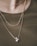 A close-up view of a person wearing multiple delicate necklaces. The jewelry consists of thin gold chains, featuring a detailed leaf-like pattern and small round pendants. The background is a green fabric or clothing, which complements the gold tones of the necklaces.