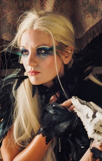 A woman with striking makeup featuring bold blue eyeshadow that extends beyond her eyelids, blonde hair, and a red lipstick. Her attire includes black feathers around her shoulders and lace gloves. She holds a white object that resembles a piece of coral or bone.