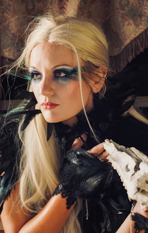 A woman with striking makeup featuring bold blue eyeshadow that extends beyond her eyelids, blonde hair, and a red lipstick. Her attire includes black feathers around her shoulders and lace gloves. She holds a white object that resembles a piece of coral or bone.