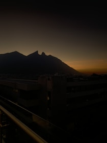 A dramatic landscape featuring a mountain range silhouetted against a twilight sky, with a cityscape in the foreground. The sky transitions from deep blue to orange near the horizon, suggesting sunset or sunrise, casting a serene and contemplative mood.