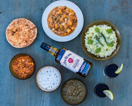 A variety of Indian dishes is arranged on a blue surface, including naan bread, a bowl of curry with paneer, a plate of rice garnished with cilantro and green chilies, several other bowls with different curry or dip, a bottle of bourbon, and two glasses with a dark beverage, each garnished with a lemon slice.