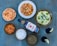 A variety of Indian dishes is arranged on a blue surface, including naan bread, a bowl of curry with paneer, a plate of rice garnished with cilantro and green chilies, several other bowls with different curry or dip, a bottle of bourbon, and two glasses with a dark beverage, each garnished with a lemon slice.
