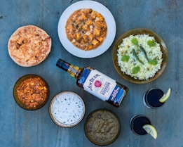 A variety of Indian dishes is arranged on a blue surface, including naan bread, a bowl of curry with paneer, a plate of rice garnished with cilantro and green chilies, several other bowls with different curry or dip, a bottle of bourbon, and two glasses with a dark beverage, each garnished with a lemon slice.