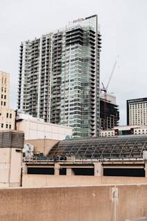 A modern cityscape featuring a tall, multi-story building with a glass facade, alongside other buildings. A construction crane is visible in the background, indicating ongoing development. The scene includes various architectural styles and a dome-shaped structure in the foreground.