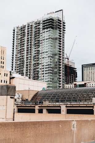 A modern cityscape featuring a tall, multi-story building with a glass facade, alongside other buildings. A construction crane is visible in the background, indicating ongoing development. The scene includes various architectural styles and a dome-shaped structure in the foreground.