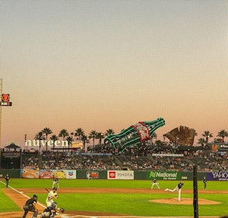 A baseball game taking place at a stadium during sunset, with a large crowd of spectators in the stands. The outfield features well-lit advertisements and a giant Coca-Cola bottle and baseball glove. The sky is clear and has a warm hue, with palm trees silhouetted against it.