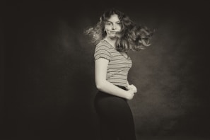 A monochrome portrait of a woman with curly hair in motion, wearing a striped t-shirt. She is positioned slightly to the side against a plain, dark backdrop, with an expression that suggests confidence.