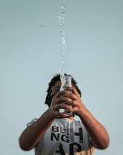 A person holding a clear glass bottle with water spraying upwards in a dynamic motion against a pale blue sky. The person is wearing a light-colored T-shirt with bold black letters and glasses, looking upwards towards the water.