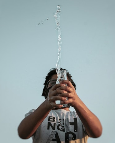 A person holding a clear glass bottle with water spraying upwards in a dynamic motion against a pale blue sky. The person is wearing a light-colored T-shirt with bold black letters and glasses, looking upwards towards the water.
