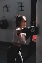 A young woman is practicing boxing with a heavy punching bag indoors. She wears a white T-shirt, black leggings, and boxing gloves. The light casts dramatic shadows, highlighting her focused expression. In the background, gym equipment is visible.