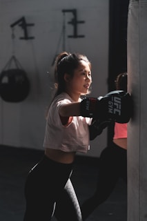 A young woman is practicing boxing with a heavy punching bag indoors. She wears a white T-shirt, black leggings, and boxing gloves. The light casts dramatic shadows, highlighting her focused expression. In the background, gym equipment is visible.