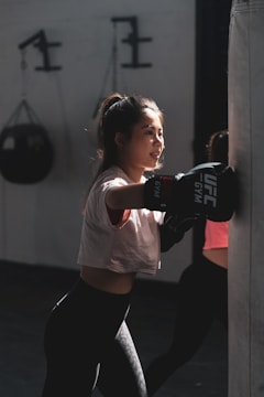 A young woman is practicing boxing with a heavy punching bag indoors. She wears a white T-shirt, black leggings, and boxing gloves. The light casts dramatic shadows, highlighting her focused expression. In the background, gym equipment is visible.