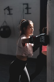 A young woman is practicing boxing with a heavy punching bag indoors. She wears a white T-shirt, black leggings, and boxing gloves. The light casts dramatic shadows, highlighting her focused expression. In the background, gym equipment is visible.
