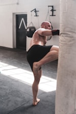 A man is engaged in a kickboxing workout, kicking a white punching bag in a gym setting. He is wearing black shorts and protective gloves, with his focus on exercise. The gym is lit with natural light from windows, illuminating the boxing equipment and mat.
