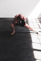 A bald individual is training with boxing gloves in a well-lit room, crouched low in a dynamic stance with one leg extended. The floor is covered with dark mats, and sunlight streams in through large windows, casting bright patterns across the room.
