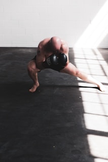 A bald individual is training with boxing gloves in a well-lit room, crouched low in a dynamic stance with one leg extended. The floor is covered with dark mats, and sunlight streams in through large windows, casting bright patterns across the room.