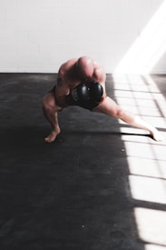 A bald individual is training with boxing gloves in a well-lit room, crouched low in a dynamic stance with one leg extended. The floor is covered with dark mats, and sunlight streams in through large windows, casting bright patterns across the room.