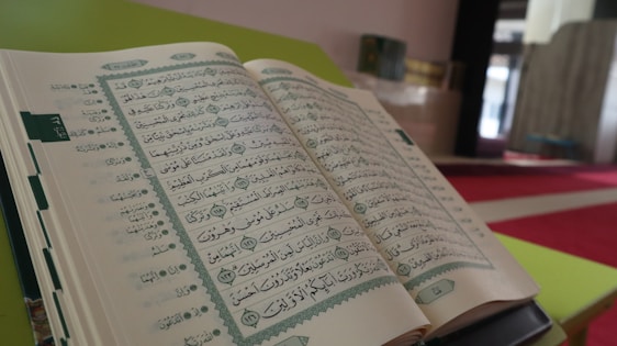 An open book displaying Arabic script is placed on a stand in a quiet indoor setting with a red and green color scheme. The background shows a mosque interior with various architectural elements blurred out, creating a sense of depth.