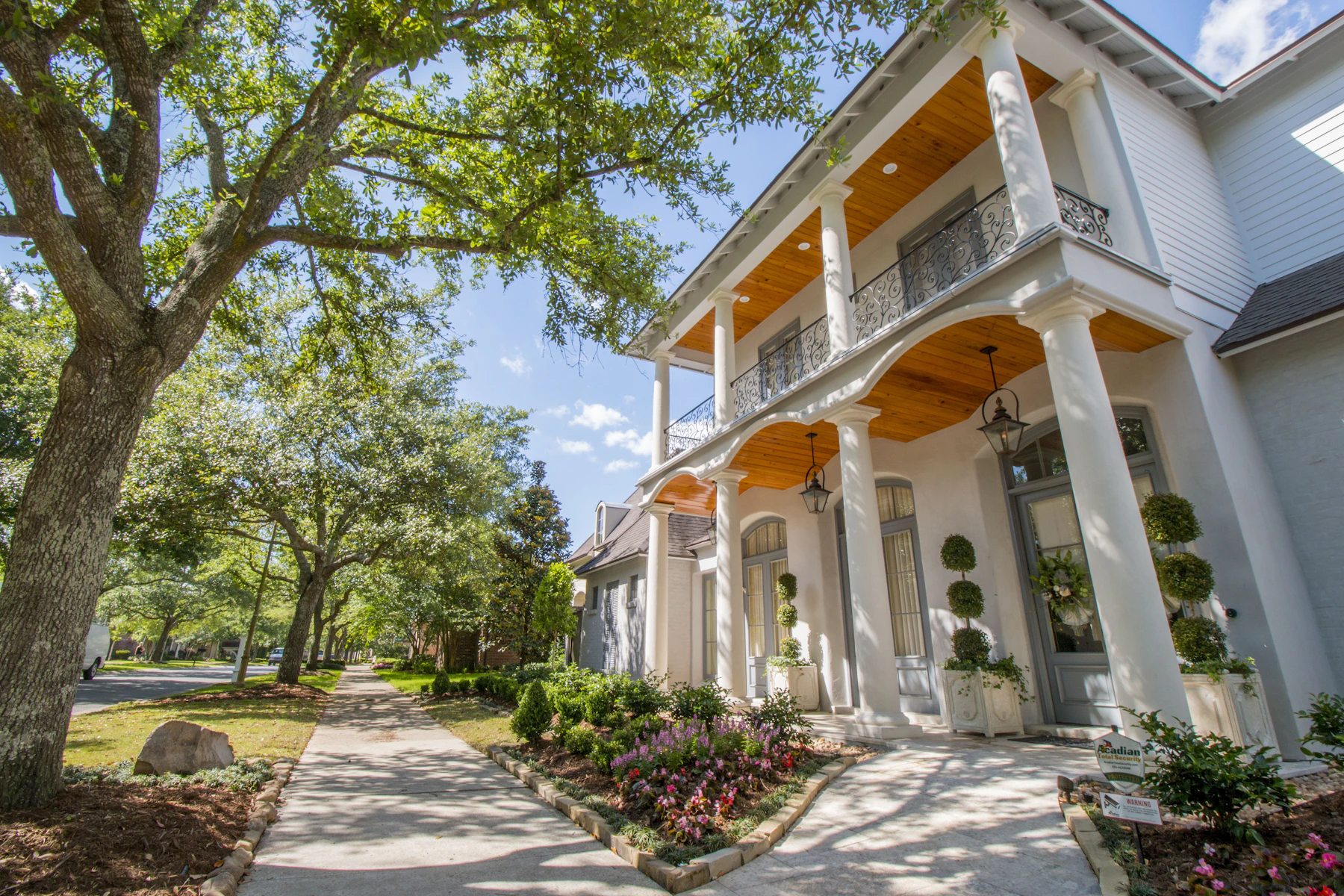 Tree-lined sidewalk beside a white two-story home with a columned porch in Lafayette, Louisiana.