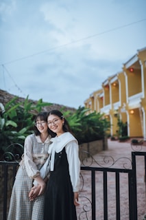 Two young women are standing close together near a decorative black metal fence, smiling warmly. They are both wearing stylish dresses, one in a plaid pattern and the other in a black dress with a white collar. In the background, a quaint yellow building with white trim and lush green foliage is visible under a cloudy sky.