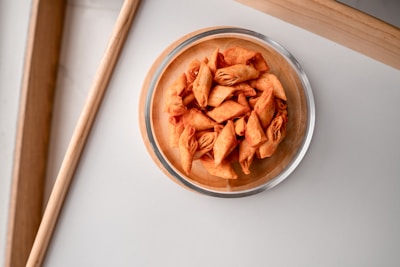 A glass bowl filled with crispy, golden-brown, folded snacks placed on a light, textured surface. The bowl is positioned near a wooden frame, adding a touch of rustic warmth to the setting.