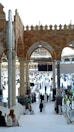 Many people dressed in religious attire gather around a large cubic structure in a grand open area with arched architecture and pillars. The space is bustling with activity, as individuals walk, sit, and observe the scene surrounded by ornate tile work and a bright, open sky.
