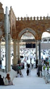 Many people dressed in religious attire gather around a large cubic structure in a grand open area with arched architecture and pillars. The space is bustling with activity, as individuals walk, sit, and observe the scene surrounded by ornate tile work and a bright, open sky.