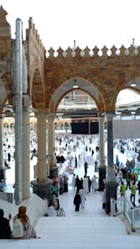 Many people dressed in religious attire gather around a large cubic structure in a grand open area with arched architecture and pillars. The space is bustling with activity, as individuals walk, sit, and observe the scene surrounded by ornate tile work and a bright, open sky.
