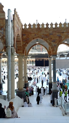 Many people dressed in religious attire gather around a large cubic structure in a grand open area with arched architecture and pillars. The space is bustling with activity, as individuals walk, sit, and observe the scene surrounded by ornate tile work and a bright, open sky.