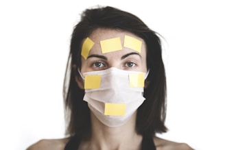 A person wearing a white face mask with multiple yellow sticky notes placed on their forehead and over the mask. The background is plain white, emphasizing the subject's expressionless face.